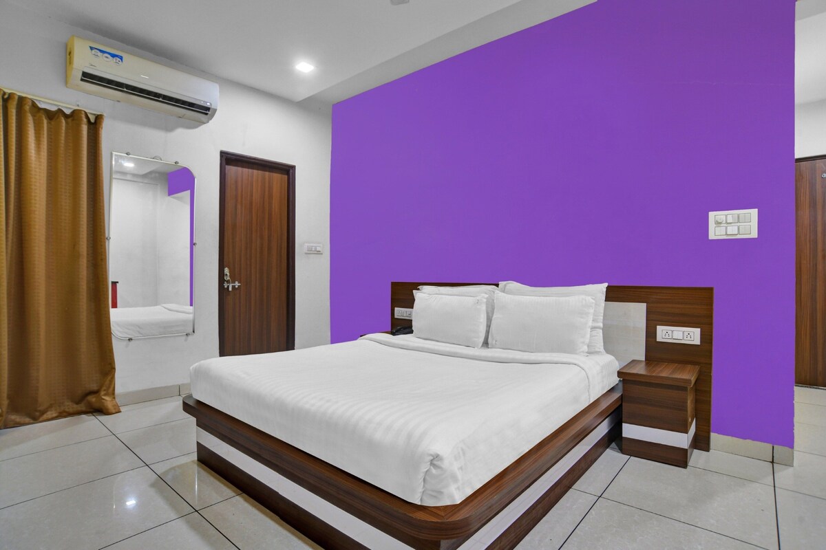 A hotel room with a bed, a nightstand, a wooden door, a bathroom with a mirror, and an air conditioning unit.