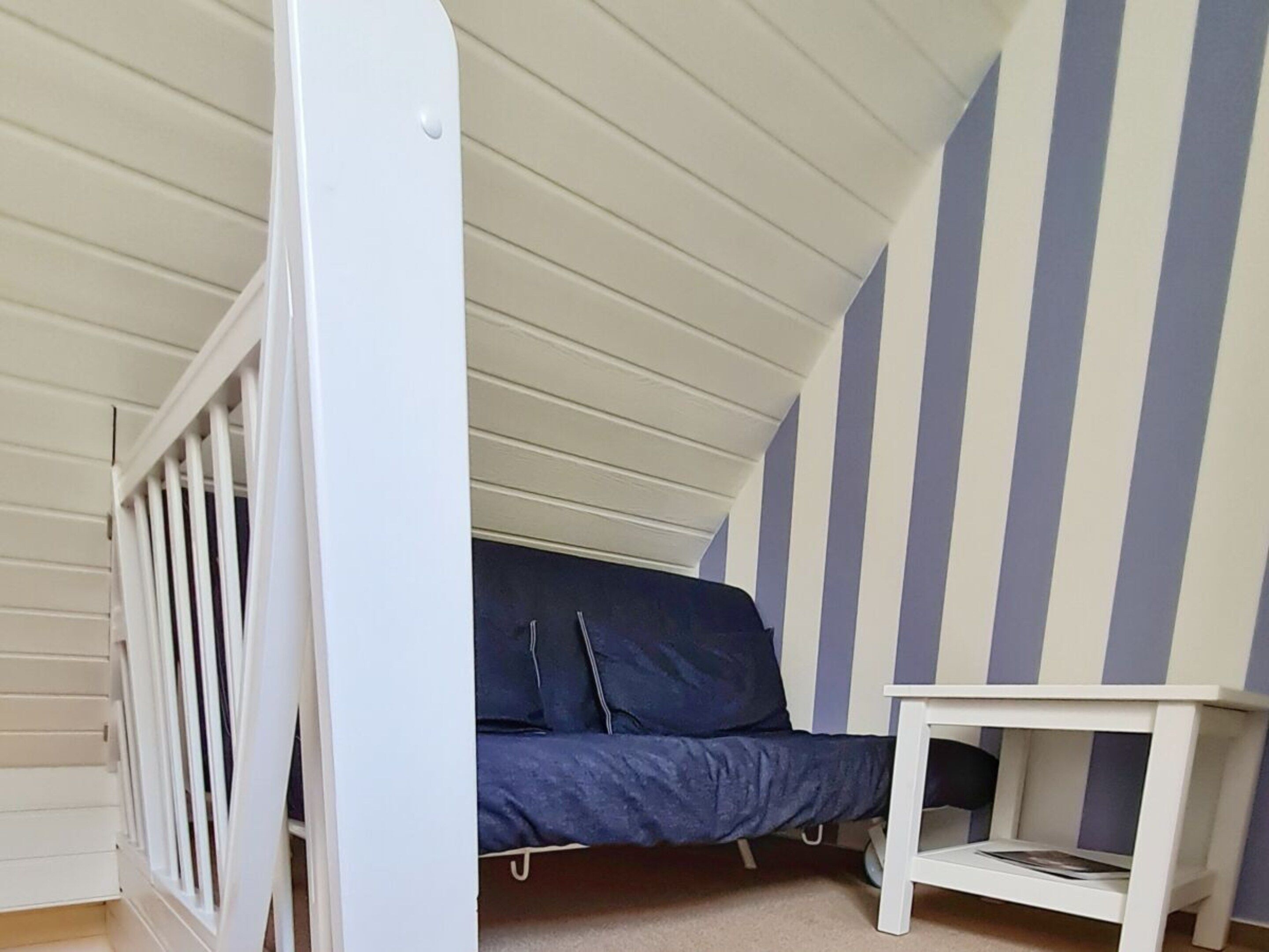 Een kamer met een schuine kap, een bed met donkerblauwe matras, een wit ledikant en een klein wit tafeltje.
