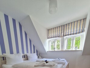 Quarto com teto inclinado, cama com roupa de cama branca, janela com valance listrado e vista para o verde lá fora.