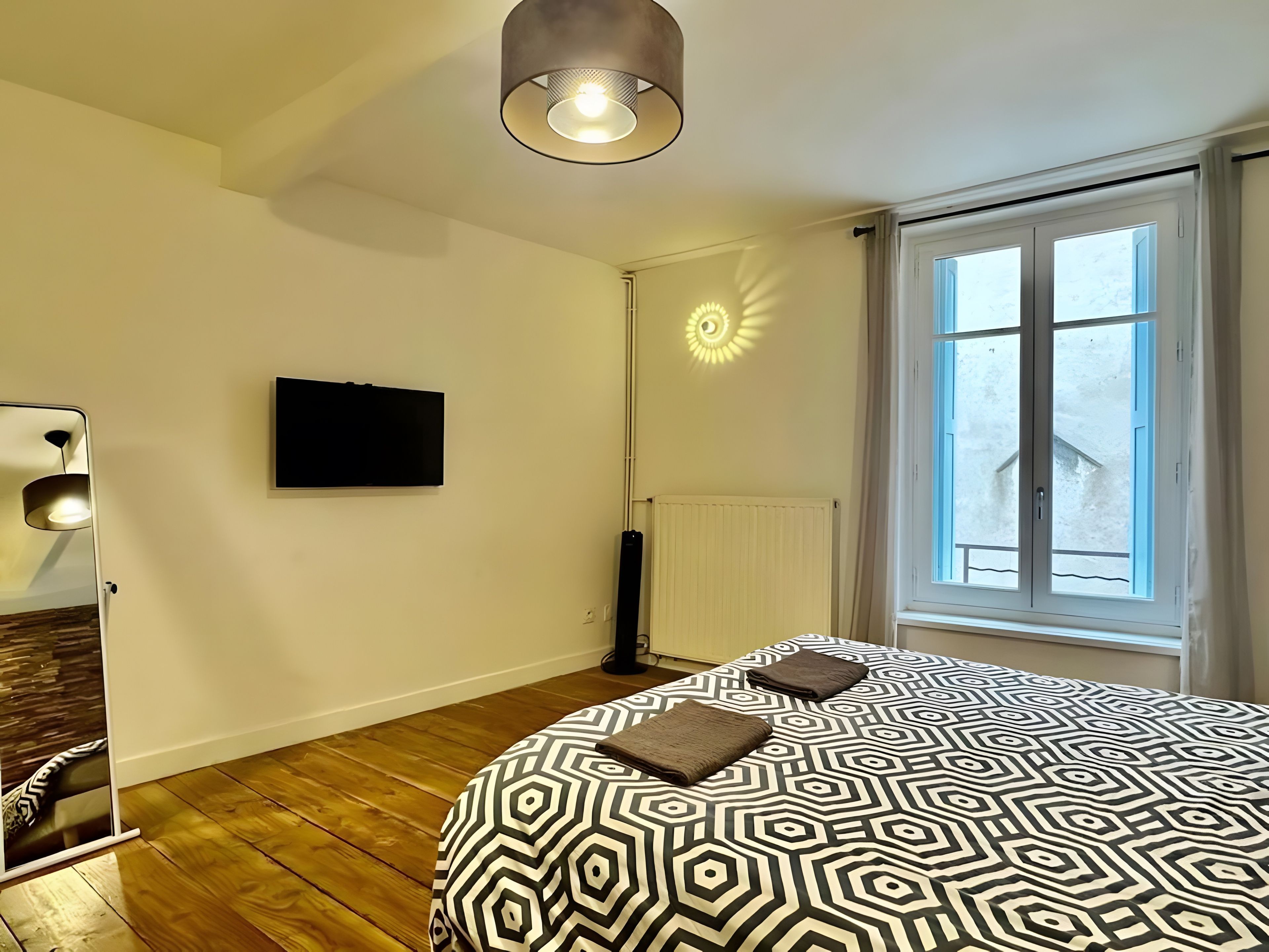 A bedroom with a patterned bedspread, a television on the wall, a standing lamp, and a window with curtains.