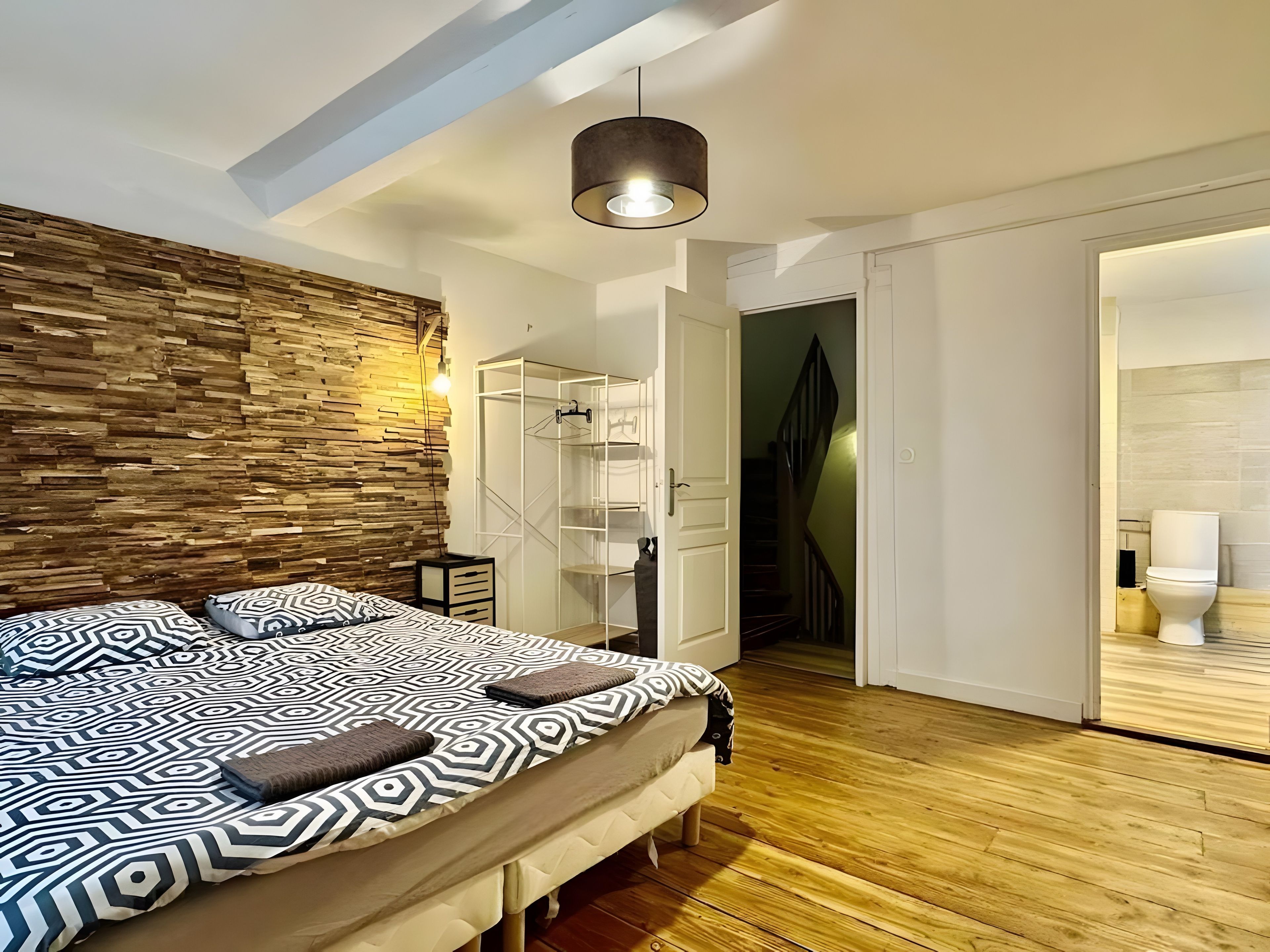 A bedroom with a patterned bedspread, a wooden wall, a hanging light fixture, and a closet with a mirrored door.