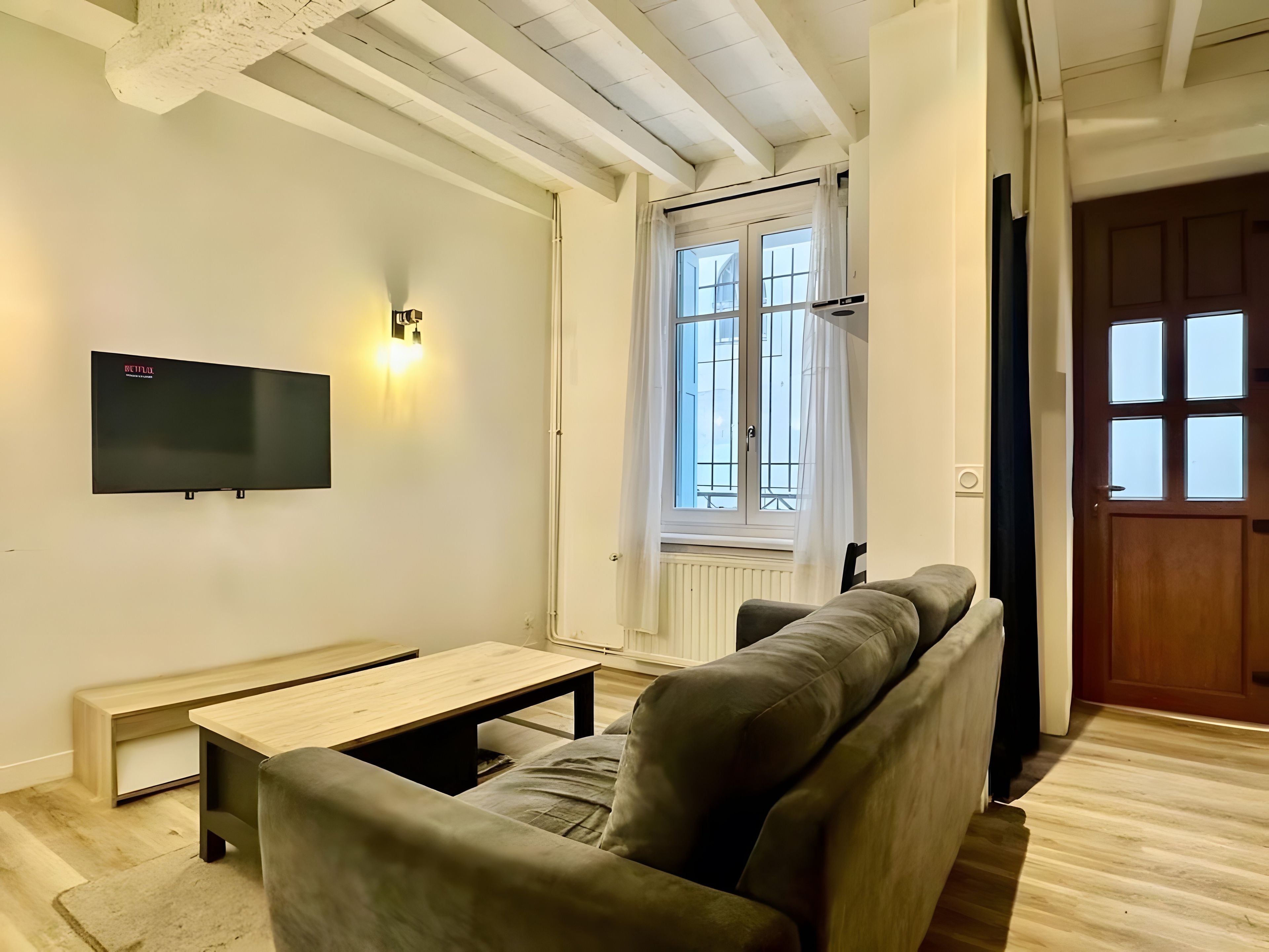 A living room with a sofa, a coffee table, a flat-screen TV mounted on the wall, and a window with curtains.