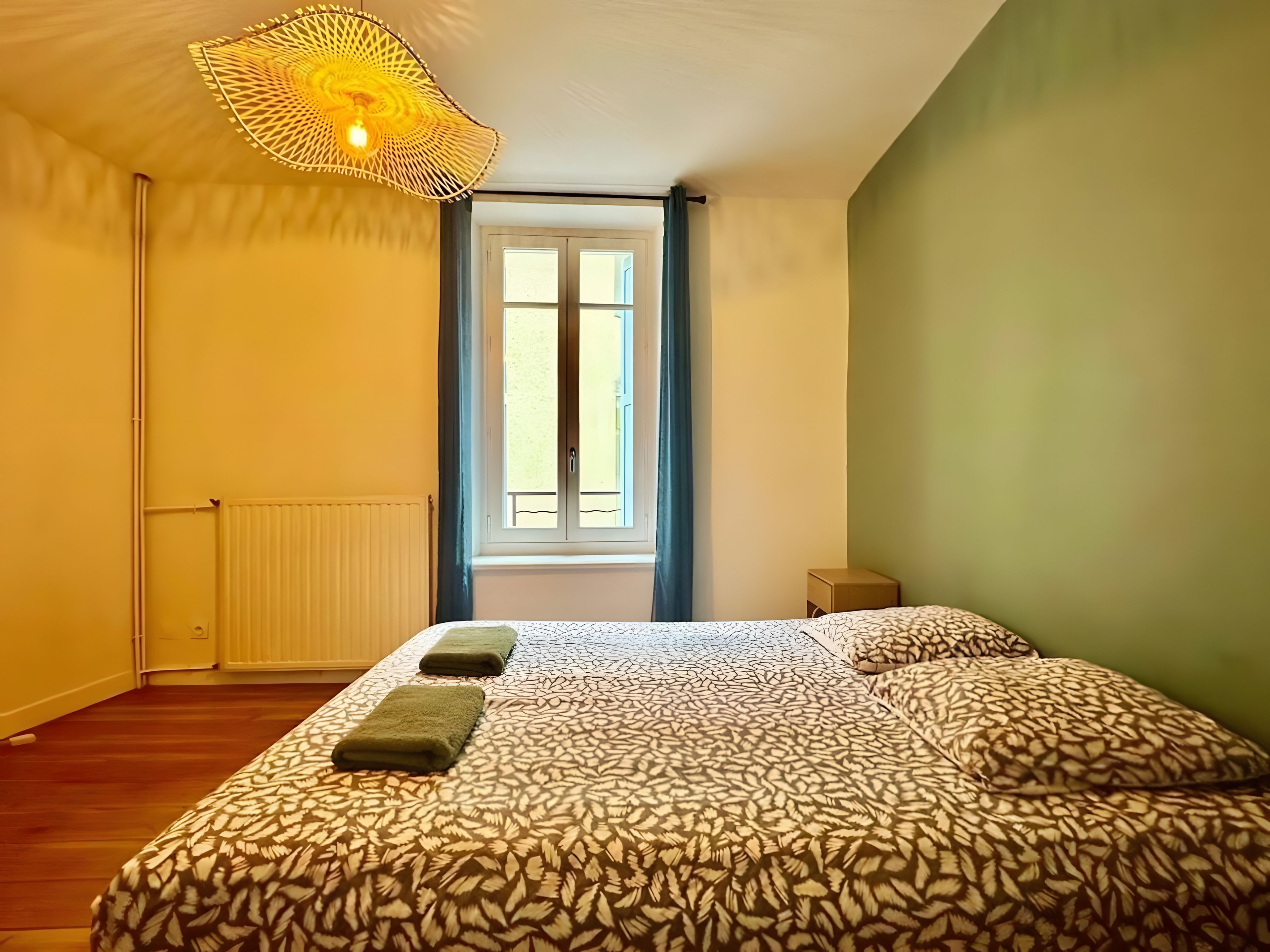 Ein Schlafzimmer mit einem gemusterten Bett, einer Hängelampe, einem Heizkörper und einem Fenster mit Vorhängen.