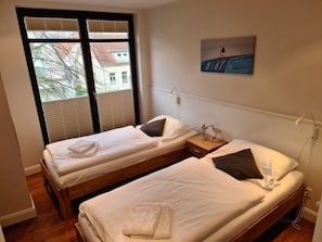 A room with two single beds, a wooden nightstand with a lamp, and a window with blinds. - Rothtraut Remise 4 (Wyk auf Foehr)