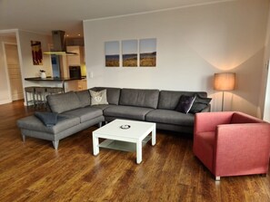 Una sala moderna con un sofá seccional gris, una mesa de centro blanca y un sillón rojo. Se ve un área de cocina al fondo.