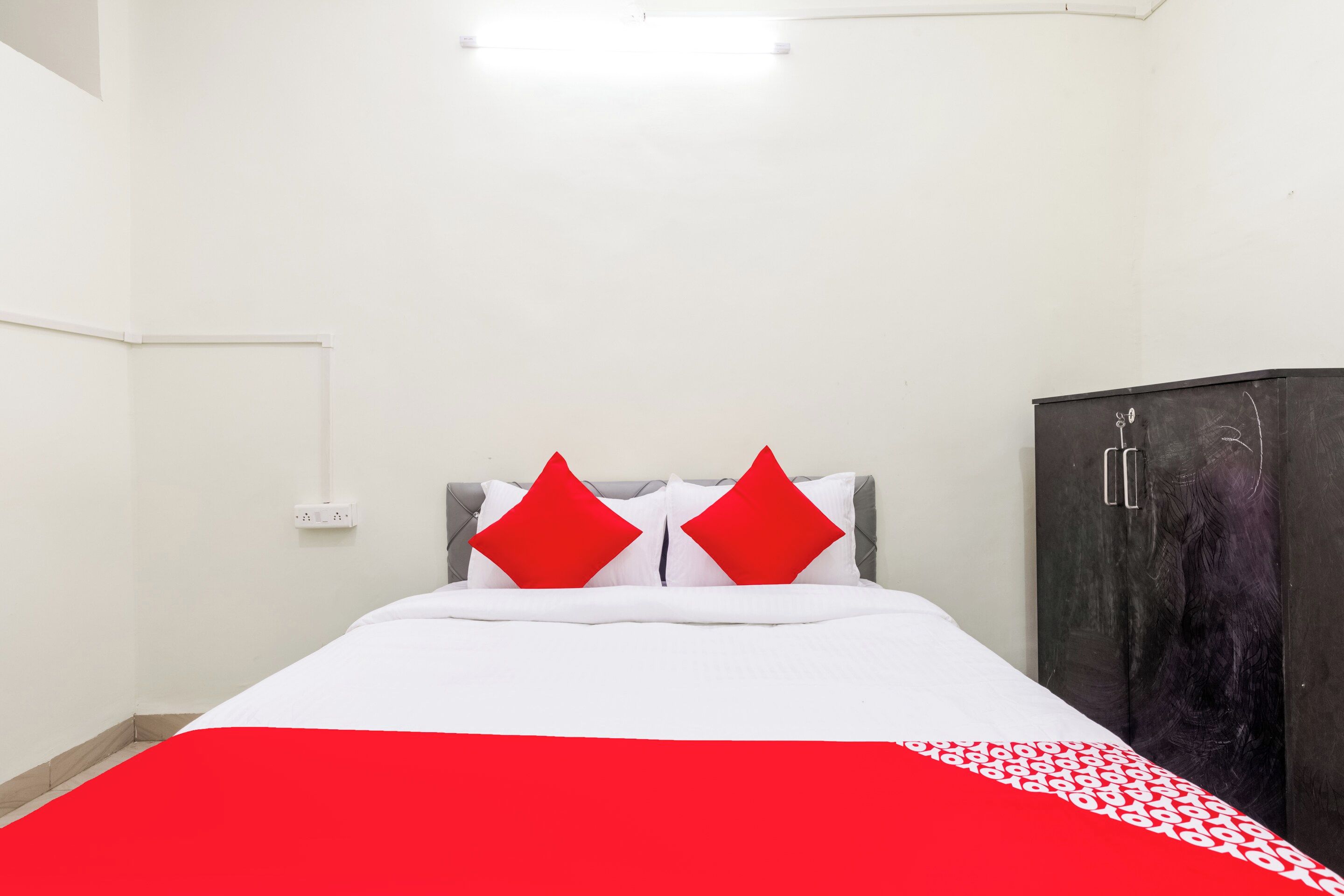 A bedroom with a bed, two red pillows, a white bedspread, a grey headboard, a dark wardrobe, and a white electrical outlet on the wall.