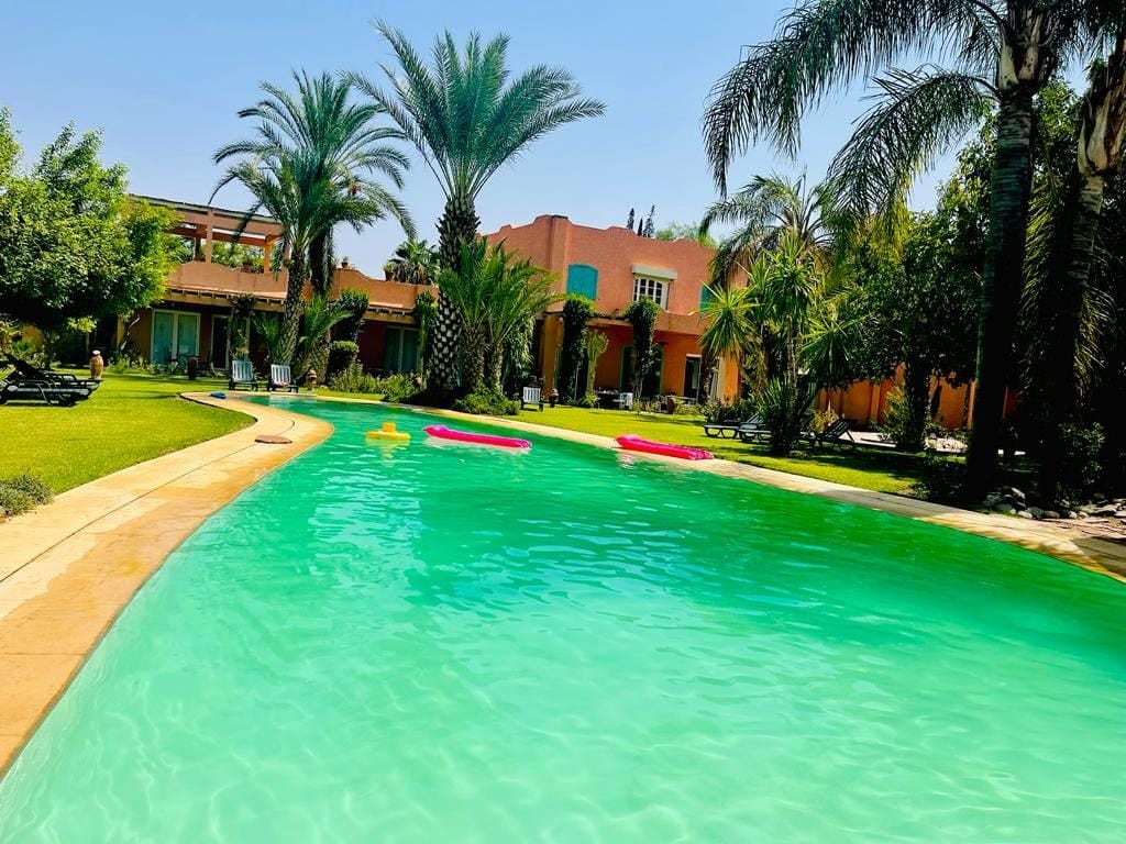 A swimming pool with clear water, surrounded by palm trees and a building with a red roof.