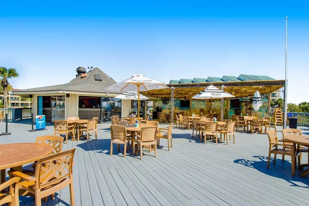 Una terrazza esterna con tavoli e sedie di legno, ombrelloni e un edificio con tetto spiovente sullo sfondo.