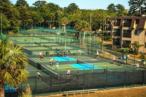 Een tenniskomplex met meerdere banen, omgeven door een hek, naast een meerlaags gebouw.