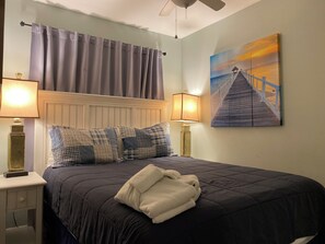 Een net opgemaakt bed met een donge deken, twee lampen aan weerszijden, een schilderij van een pier en een plafondventilator.