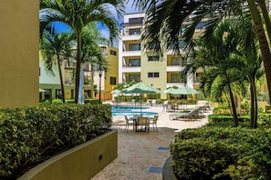 A pool area with lounge chairs and umbrellas, surrounded by palm trees and a building with balconies. - Loft-Style 2BR Condo w/ Pool View, Palm Beach (Noord)