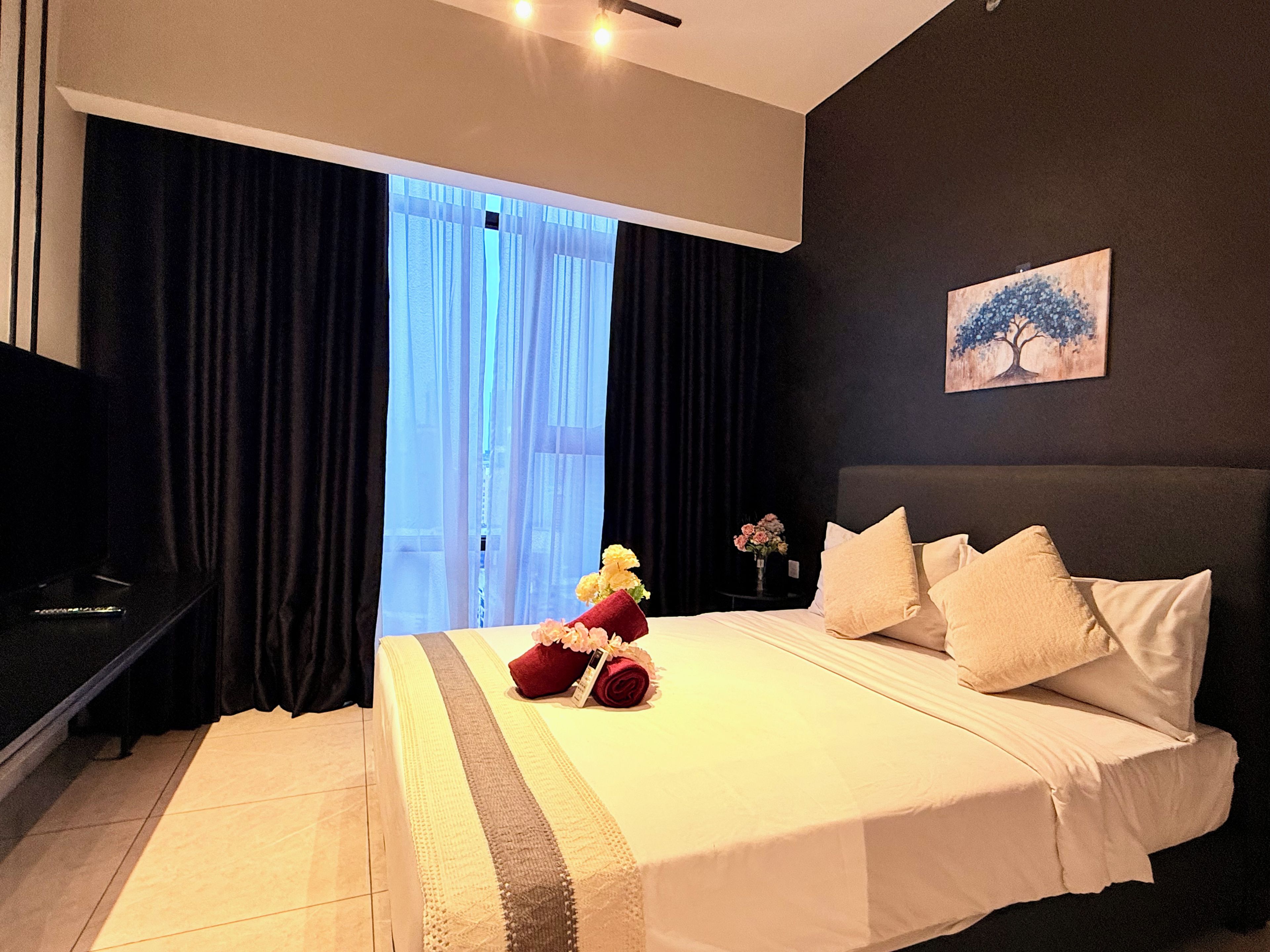 A hotel room with a large bed, a nightstand, a vase with flowers, and a painting on the wall.