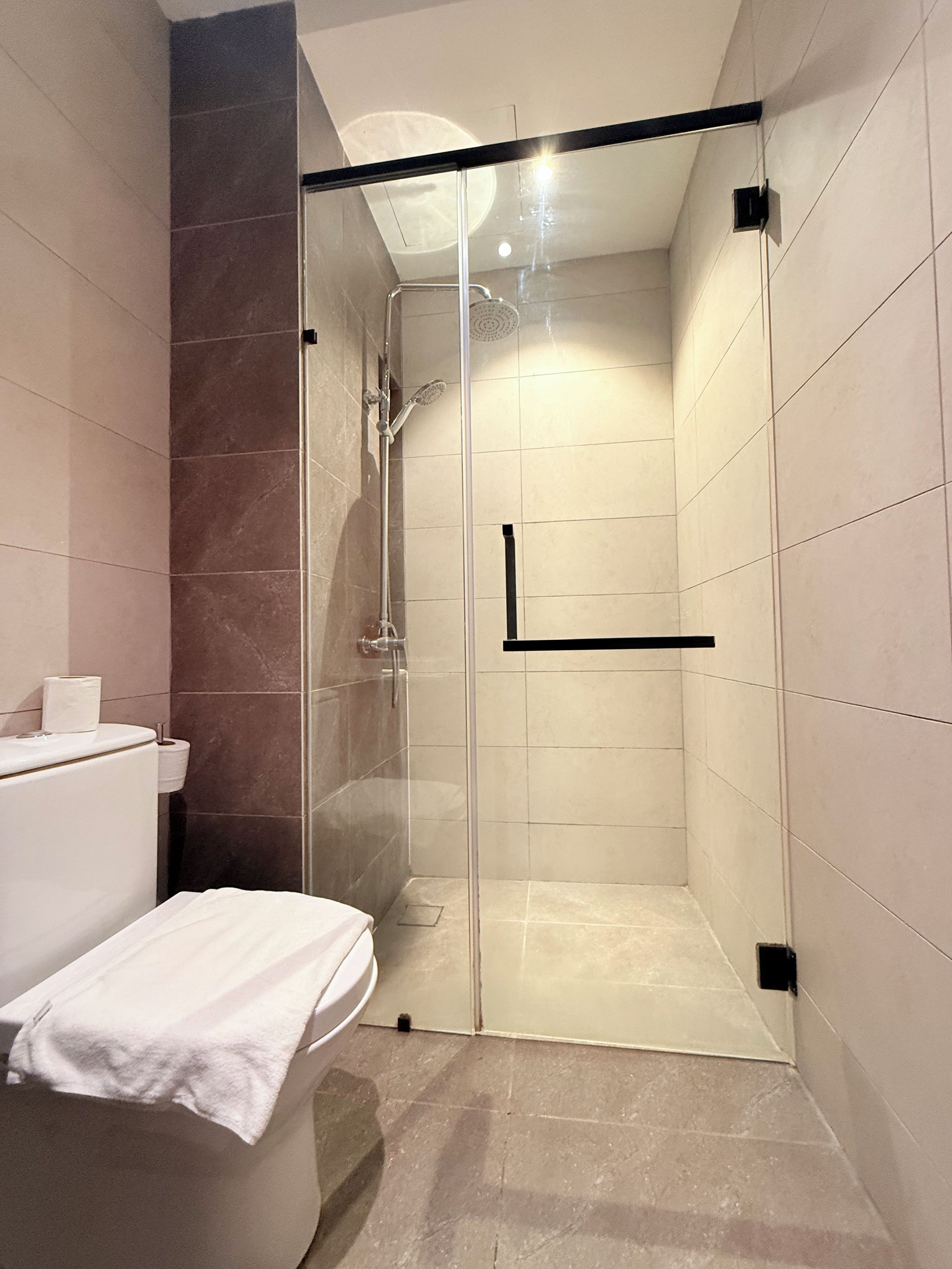 Cabina de ducha con paredes de vidrio, un cabezal de ducha manual, una toalla en el suelo y un inodoro con un rollo de papel higiénico.