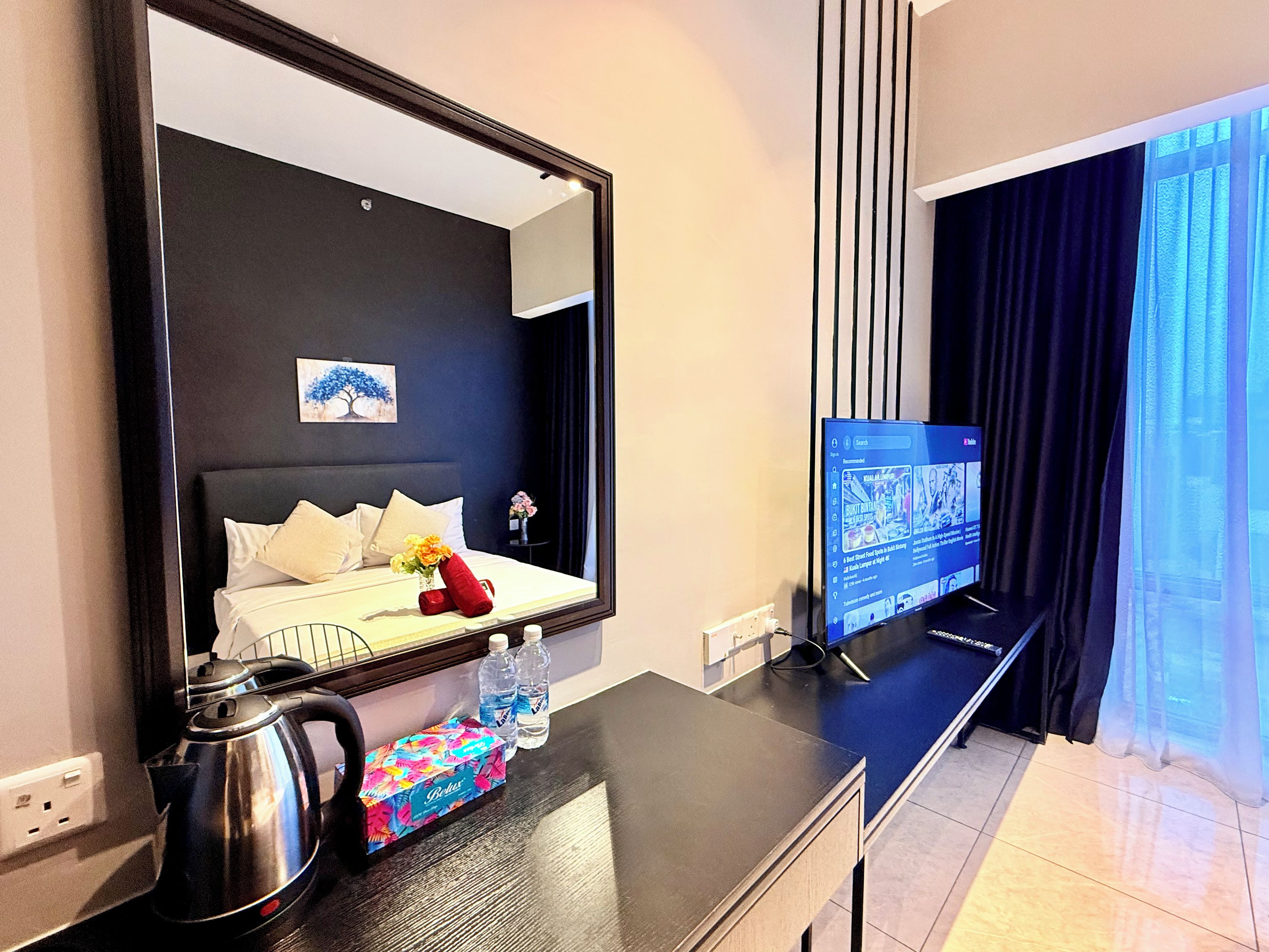 A hotel room with a bed, a TV on a stand, a kettle, a water bottle, and a small table with a box.