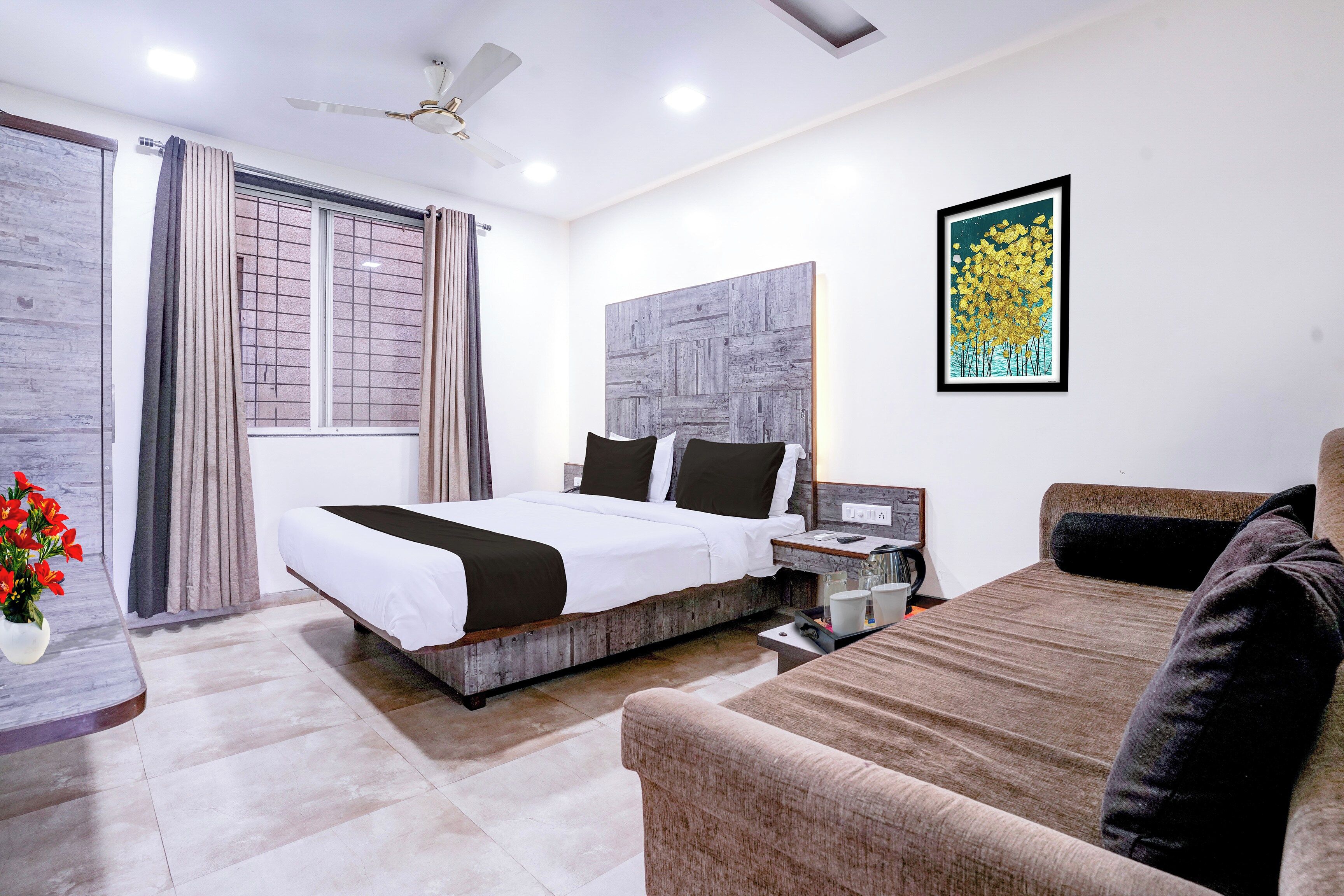 A hotel room with a bed, a sofa, a desk, a ceiling fan, a window with curtains, a wall-mounted artwork, and a vase with flowers.