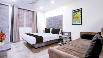 A hotel room with a bed, a sofa, a desk, a ceiling fan, a window with curtains, a wall-mounted artwork, and a vase with flowers.