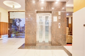 A modern elevator with a glass door, surrounded by granite walls, in a well-lit lobby area. - Townhouse by OYO Brahma Sarovar (Thanesar)