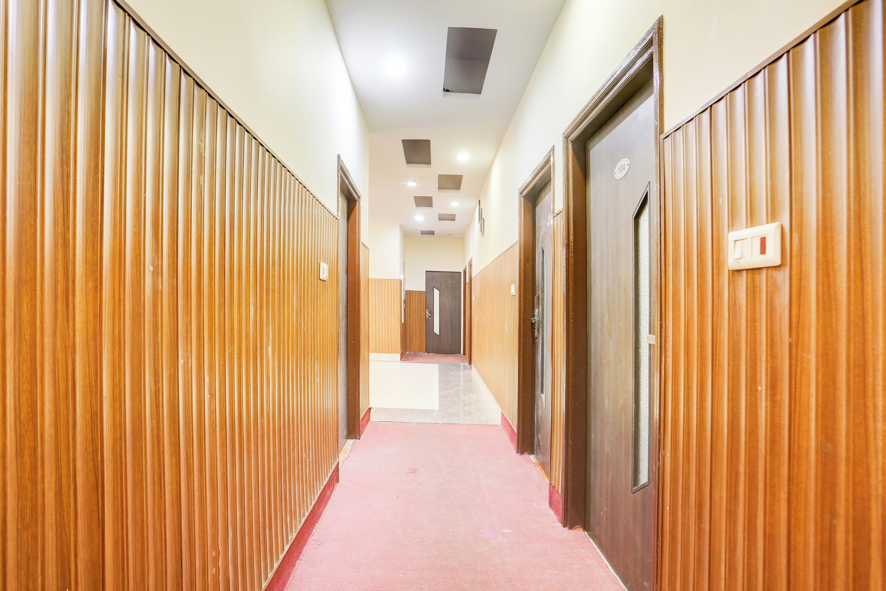 Un pasillo con paneles de madera a ambos lados, alfombra roja y puertas con números de habitación.