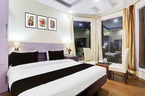 Habitación de hotel con una cama grande, un escritorio, una silla, un ventanal con cortinas y cuadros en la pared.