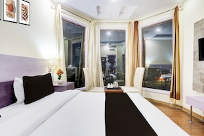 Habitación de hotel con cama, una silla, mesita de noche, lámpara, un jarrón con flores y vistas a la ciudad a través de amplias ventanas.