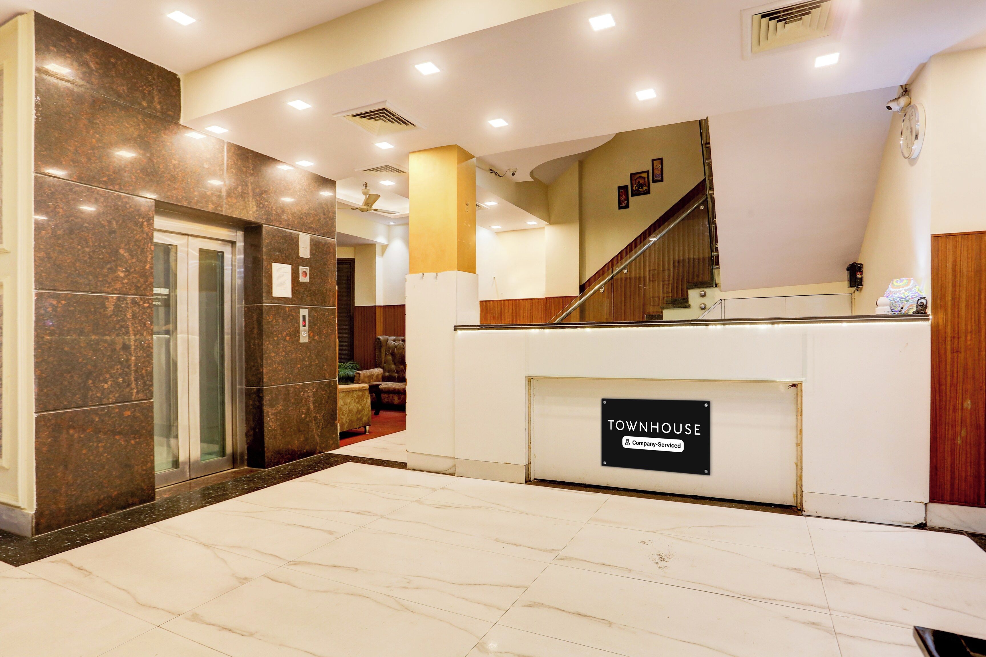 A hotel lobby with a reception desk, a staircase, and a glass elevator.