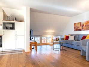 Ein geräumiger Wohnbereich mit grauem Sofa, Holzboden und einer Küche mit weißen Schränken.