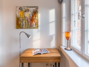 Ein Zimmer mit einem Holztisch, einer Lampe, einem Buch und einem Fenster mit Blick auf eine Ziegelwand.