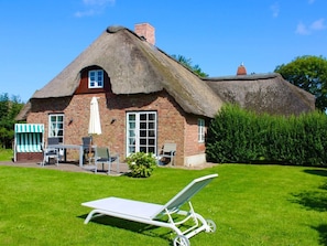 Ein Haus mit Strohdach und Backsteinfassade, eine Terrasse mit Gartenmöbeln und eine Rasenfläche.