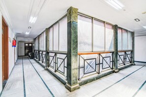 Un pasillo con una amplia estancia cerrada que cuenta con un pilar de mármol, barandillas de metal y un extintor en la pared.