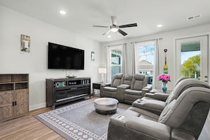 A modern living room with a flat-screen TV, a ceiling fan, and a large window with curtains. - New on Program! Beautiful 4BR Townhome in Barefoot! Close to Golf Courses! (North Myrtle Beach)