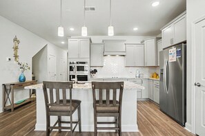 A modern kitchen with white cabinetry, a central island with two chairs, stainless steel appliances, and pendant lighting. - New on Program! Beautiful 4BR Townhome in Barefoot! Close to Golf Courses! (North Myrtle Beach)