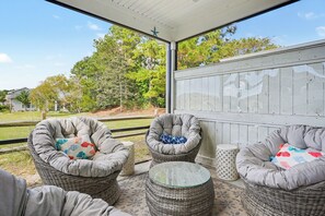 A covered outdoor seating area with wicker chairs and a glass table, overlooking a landscaped garden and trees. - New on Program! Beautiful 4BR Townhome in Barefoot! Close to Golf Courses! (North Myrtle Beach)