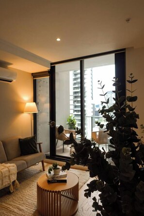 A modern living room with a sofa, a round coffee table, and a lamp. There is a large window with a view of buildings outside. - Luxury CBD Escape | Pool, Sauna & City Views (Melbourne)