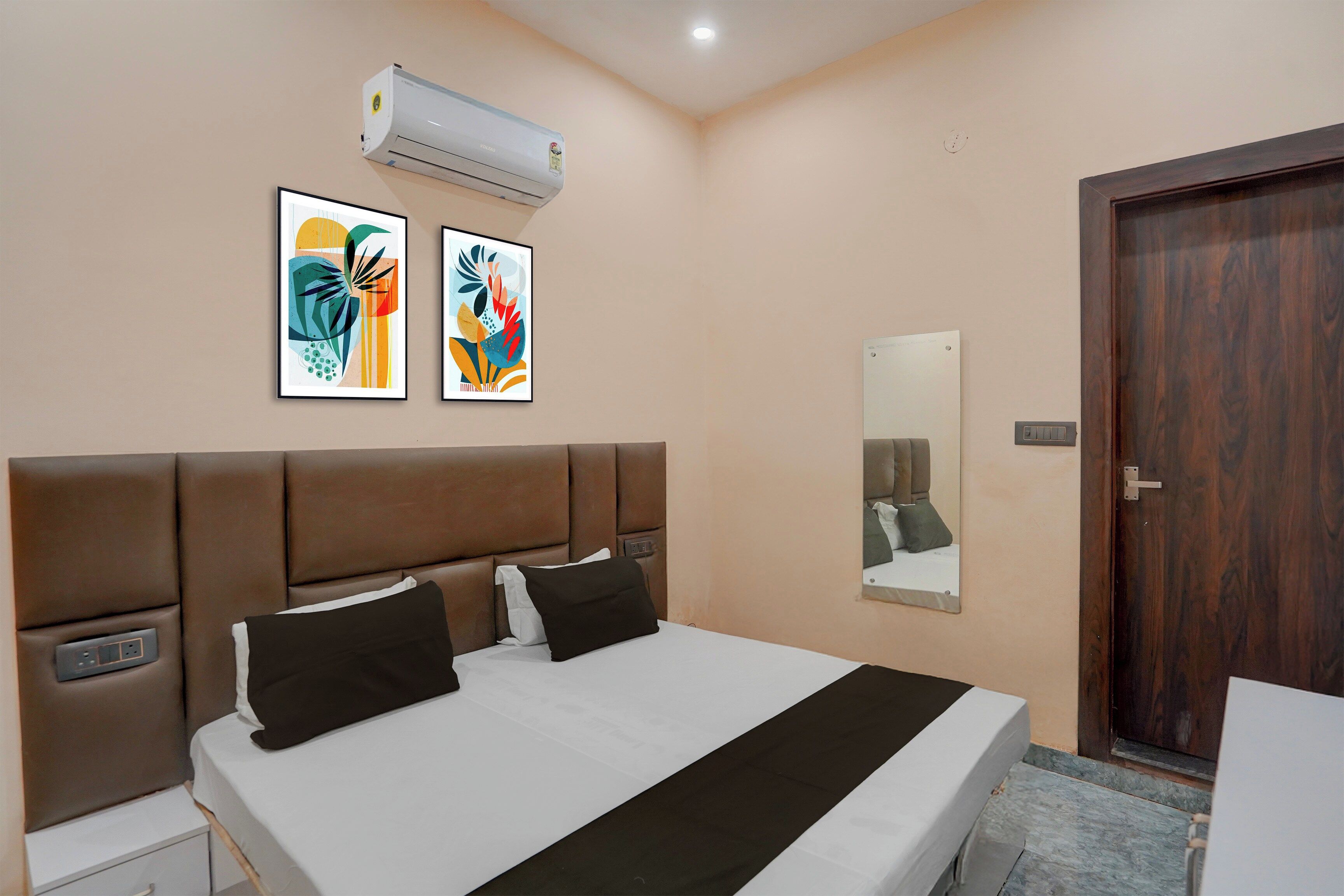 Una habitación de hotel con una cama, dos unidades de aire acondicionado fijadas a la pared, dos cuadros abstractos enmarcados, una mesita de noche con una lámpara y una puerta de madera.