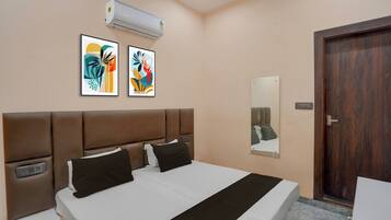 Una habitación de hotel con una cama, dos unidades de aire acondicionado fijadas a la pared, dos cuadros abstractos enmarcados, una mesita de noche con una lámpara y una puerta de madera.