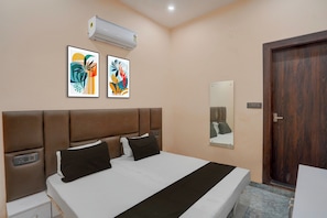 Una habitación de hotel con una cama, dos unidades de aire acondicionado fijadas a la pared, dos cuadros abstractos enmarcados, una mesita de noche con una lámpara y una puerta de madera.