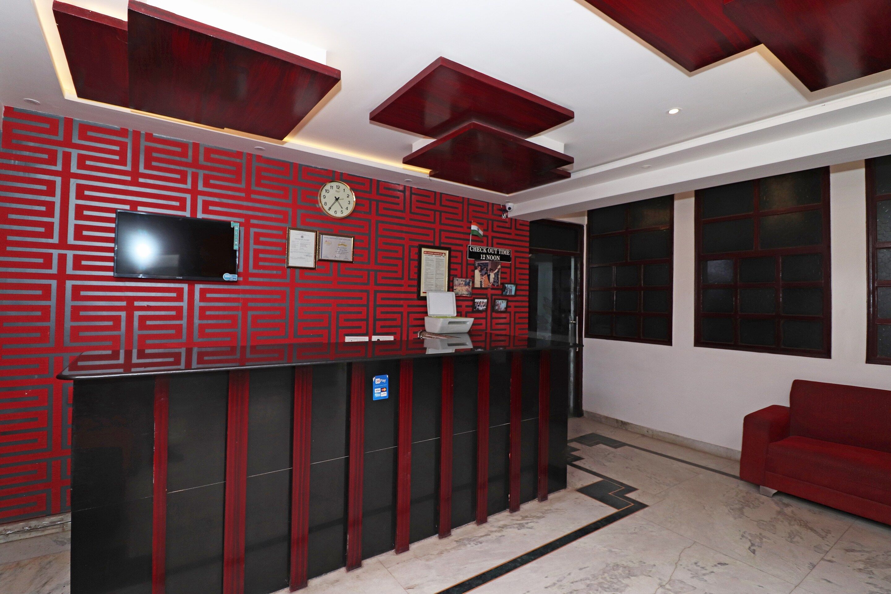 Zona de recepción con pared de estampado rojo, un reloj, una pantalla digital y un cartel de salida que indica 12 del mediodía.