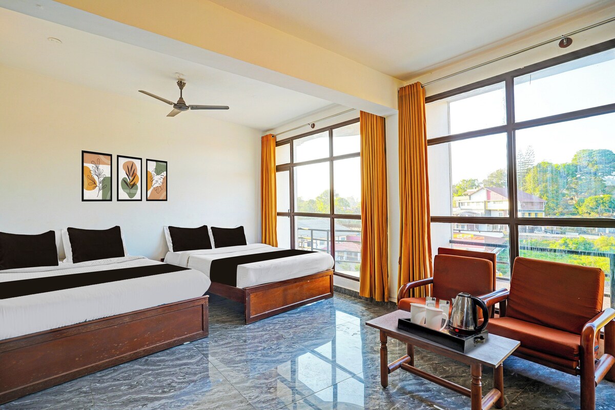 A hotel room with two beds, a table with a kettle, a chair, and large windows with curtains.