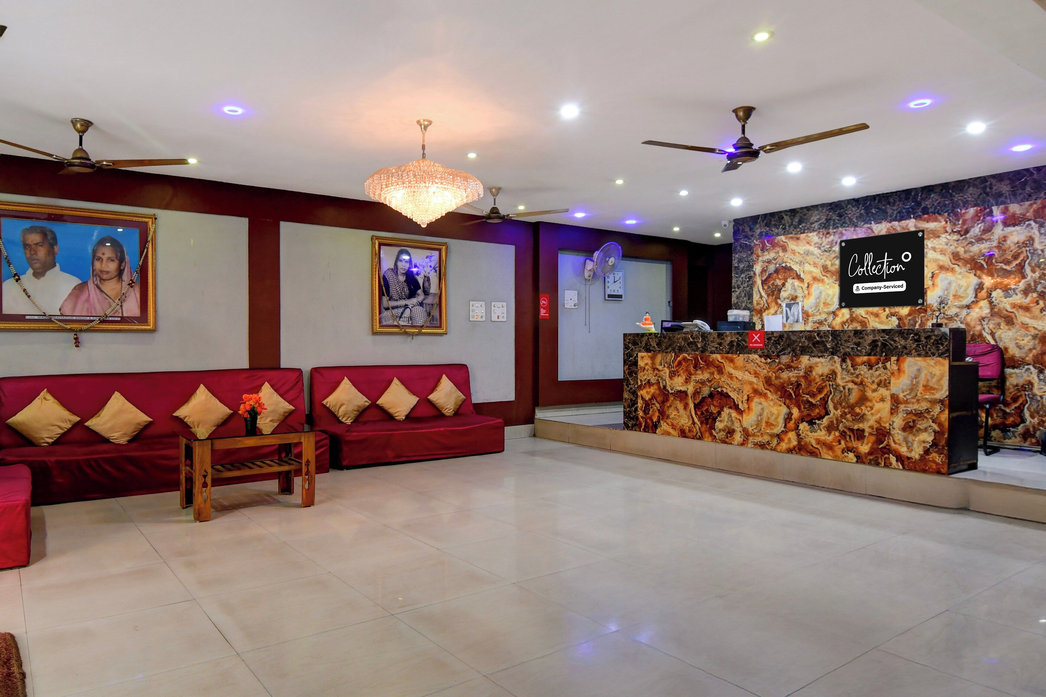 A hotel reception area with a reception desk, two red sofas with cushions, a chandelier, ceiling fans, and framed pictures on the walls.