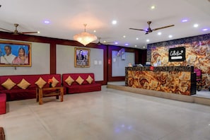 A hotel reception area with a reception desk, two red sofas with cushions, a chandelier, ceiling fans, and framed pictures on the walls.