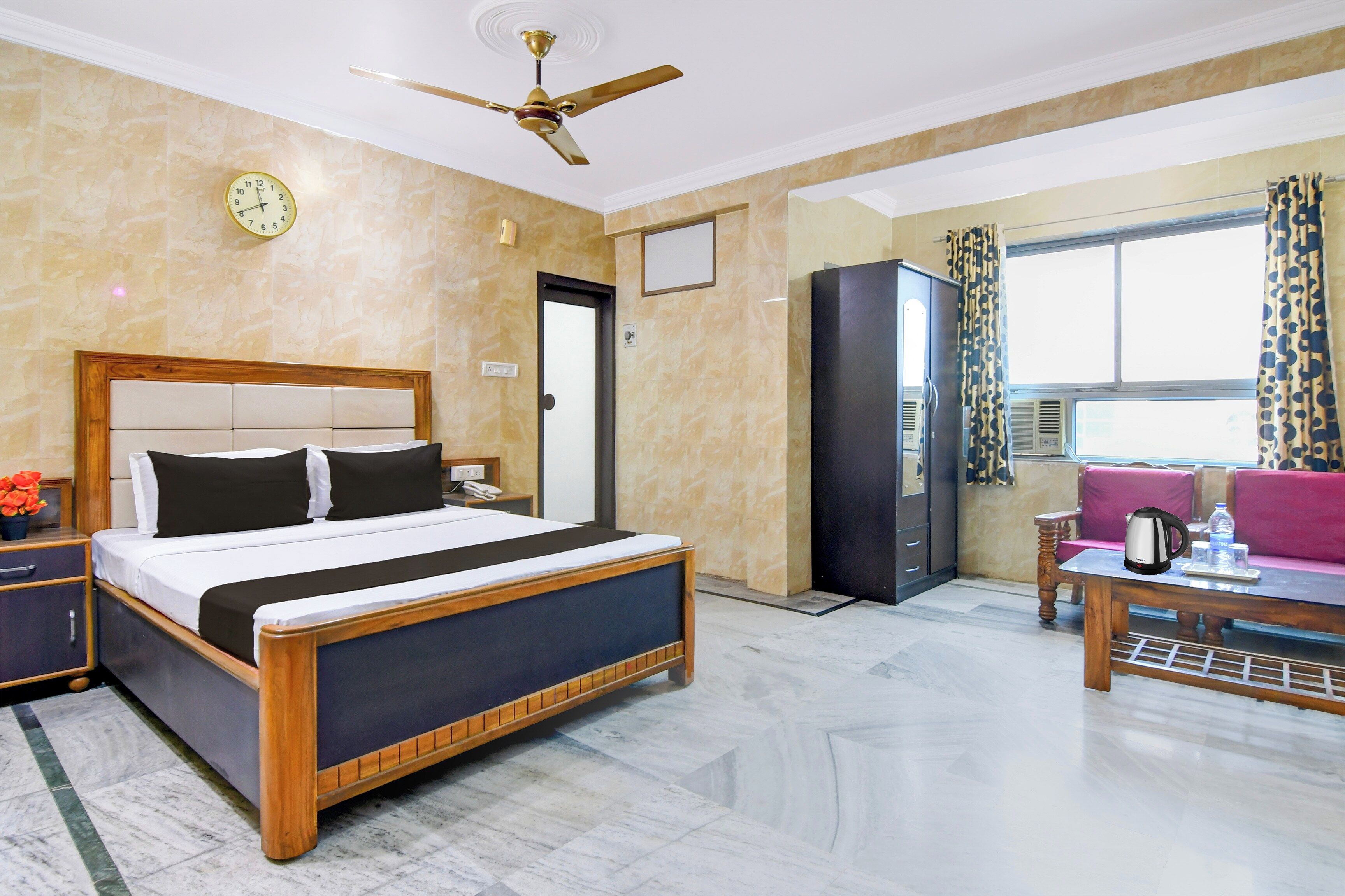 A hotel room with a bed, a desk, a chair, a wardrobe, a ceiling fan, and a window with curtains.