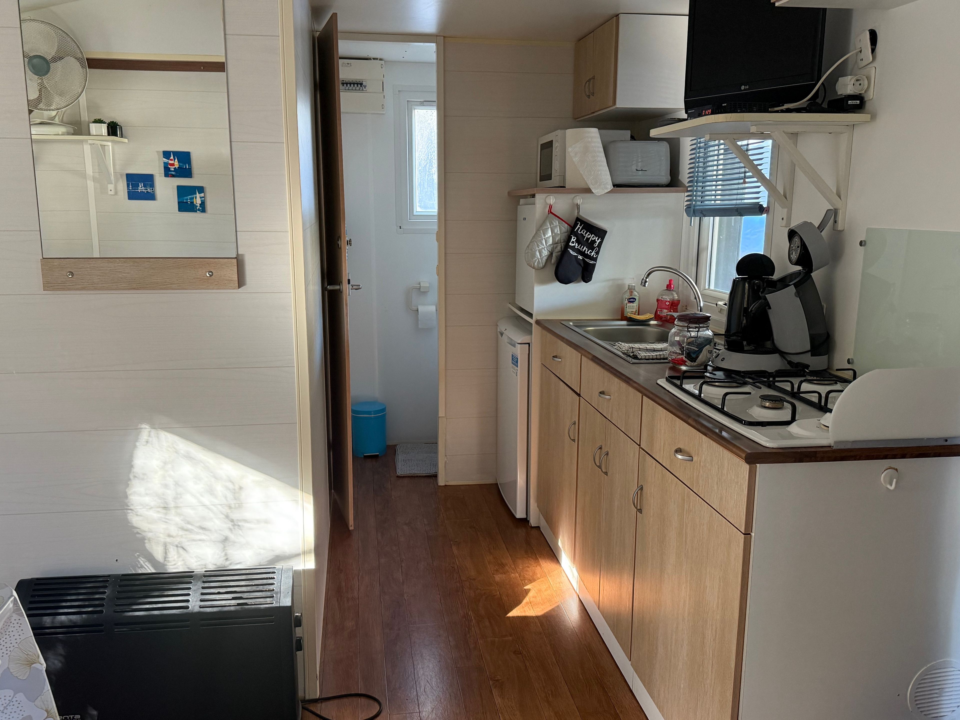Een compacte keuken met een magnetron, fornuis en gootsteen. Ook zichtbaar is een kleine prullenbak en een deur die naar een andere kamer leidt.
