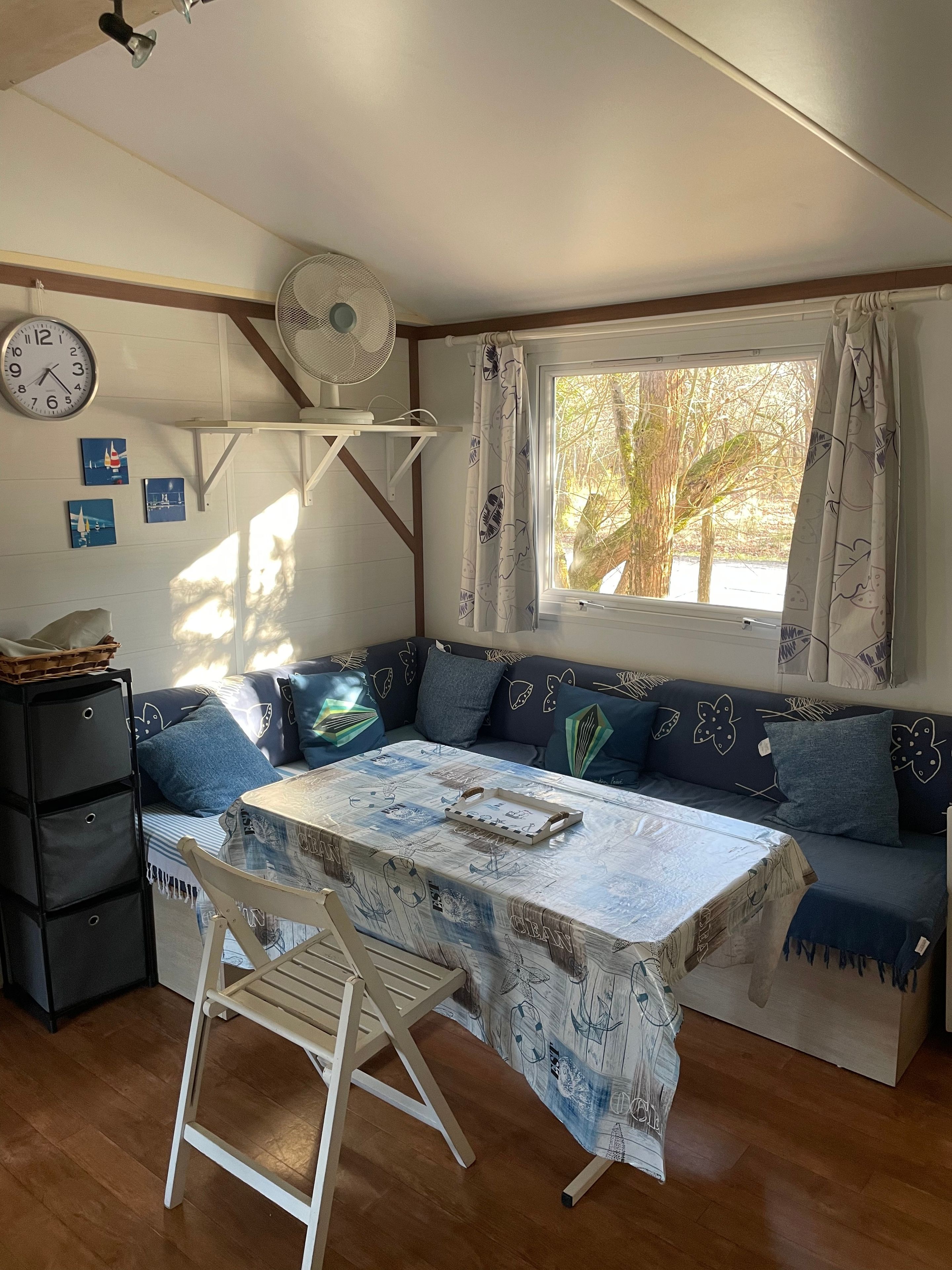 Een gezellige kamer met een bank, een tafel met tafelkleed, een ventilator, een klok en een raam met gordijnen.
