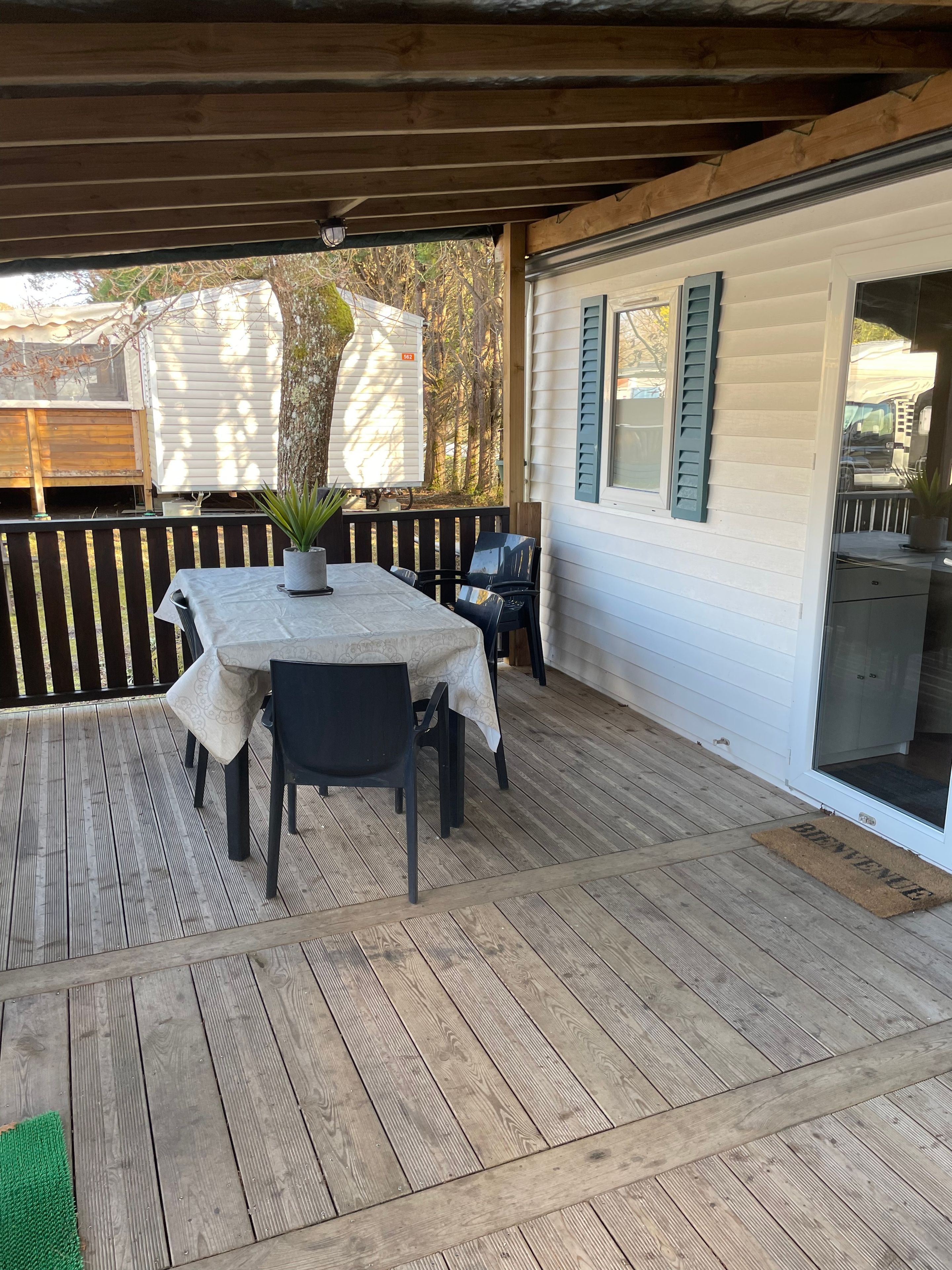 Een overdekt terras met een tafel en stoelen, een houten terras en een wit gebouw met groene luiken.