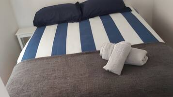Een bed met gestreept blauw-wit beddengoed, een donkerblauwe kussen en een opgerolde witte handdoek.