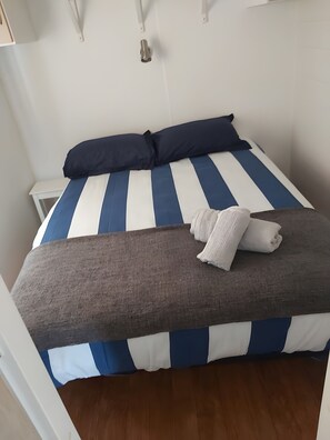 Een bed met gestreept blauw-wit beddengoed, een donkerblauwe kussen en een opgerolde witte handdoek.