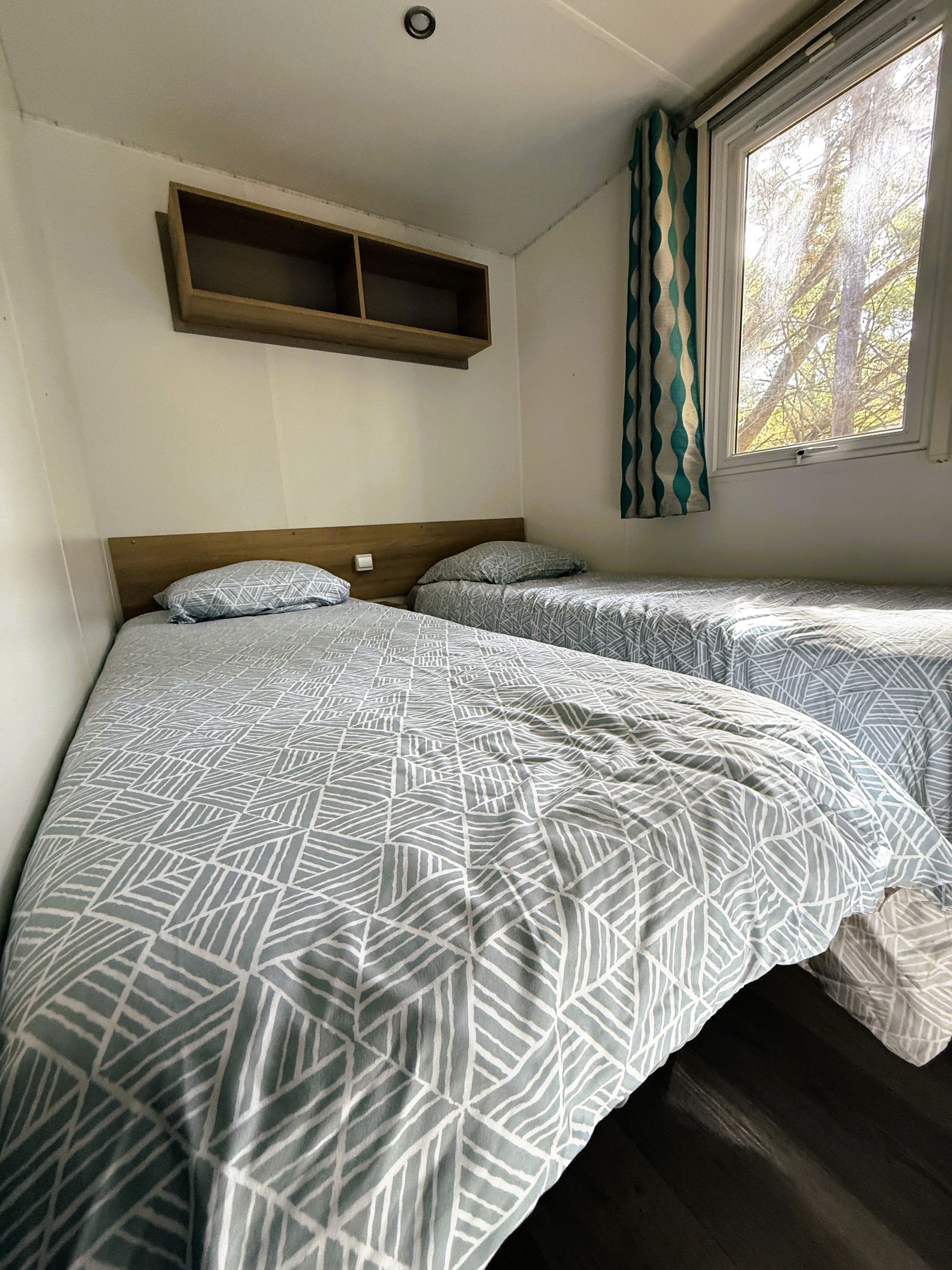 Quarto com duas camas, uma estante de madeira, janela com cortinas e uma luminária no teto.