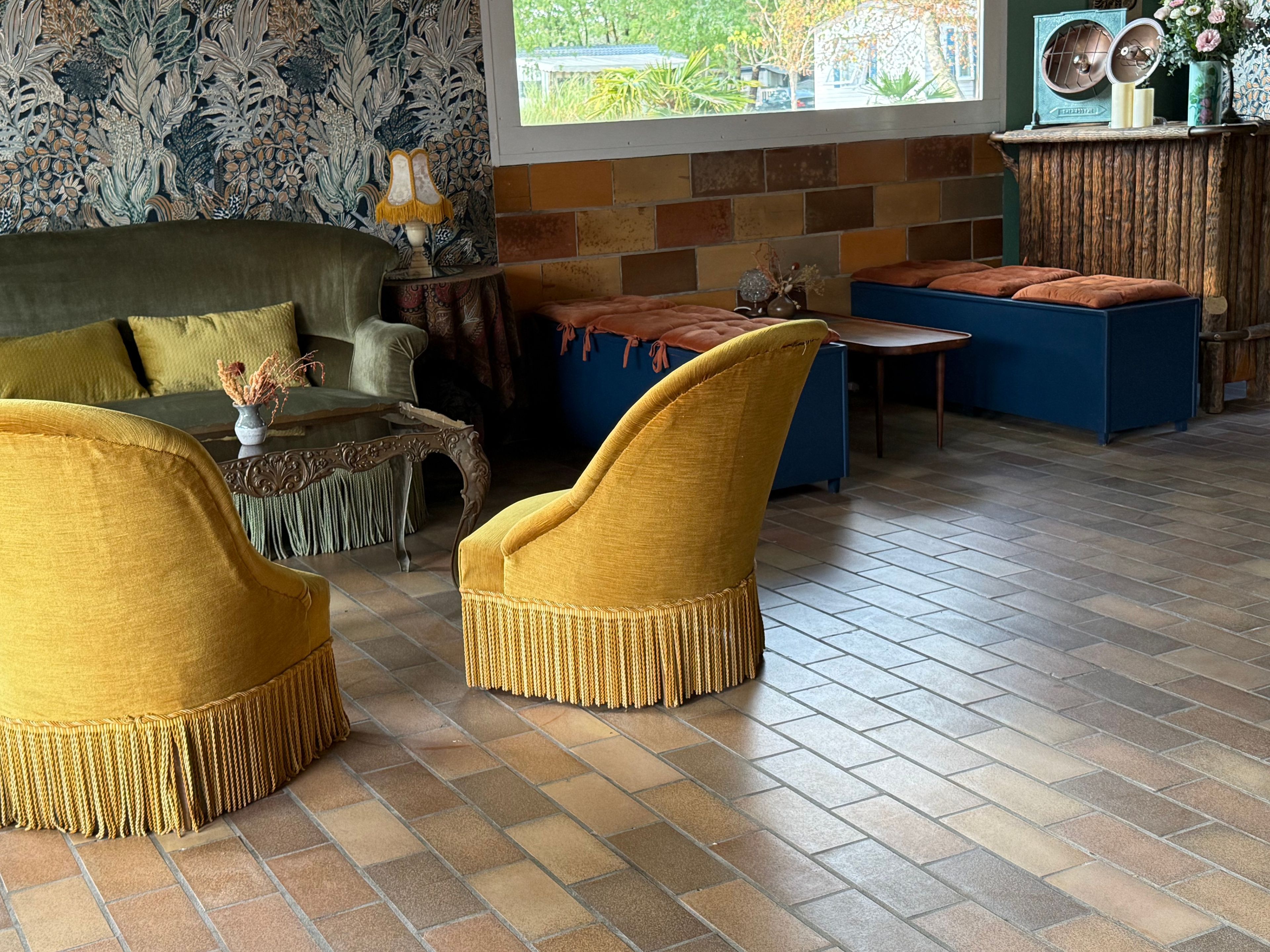 Une pièce avec un papier peint à motifs, un canapé, deux fauteuils, une petite table avec une lampe et une fenêtre donnant sur un paysage verdoyant.