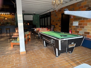 A billiards table in a room with a brick floor, a bar area with stools, and a chandelier.