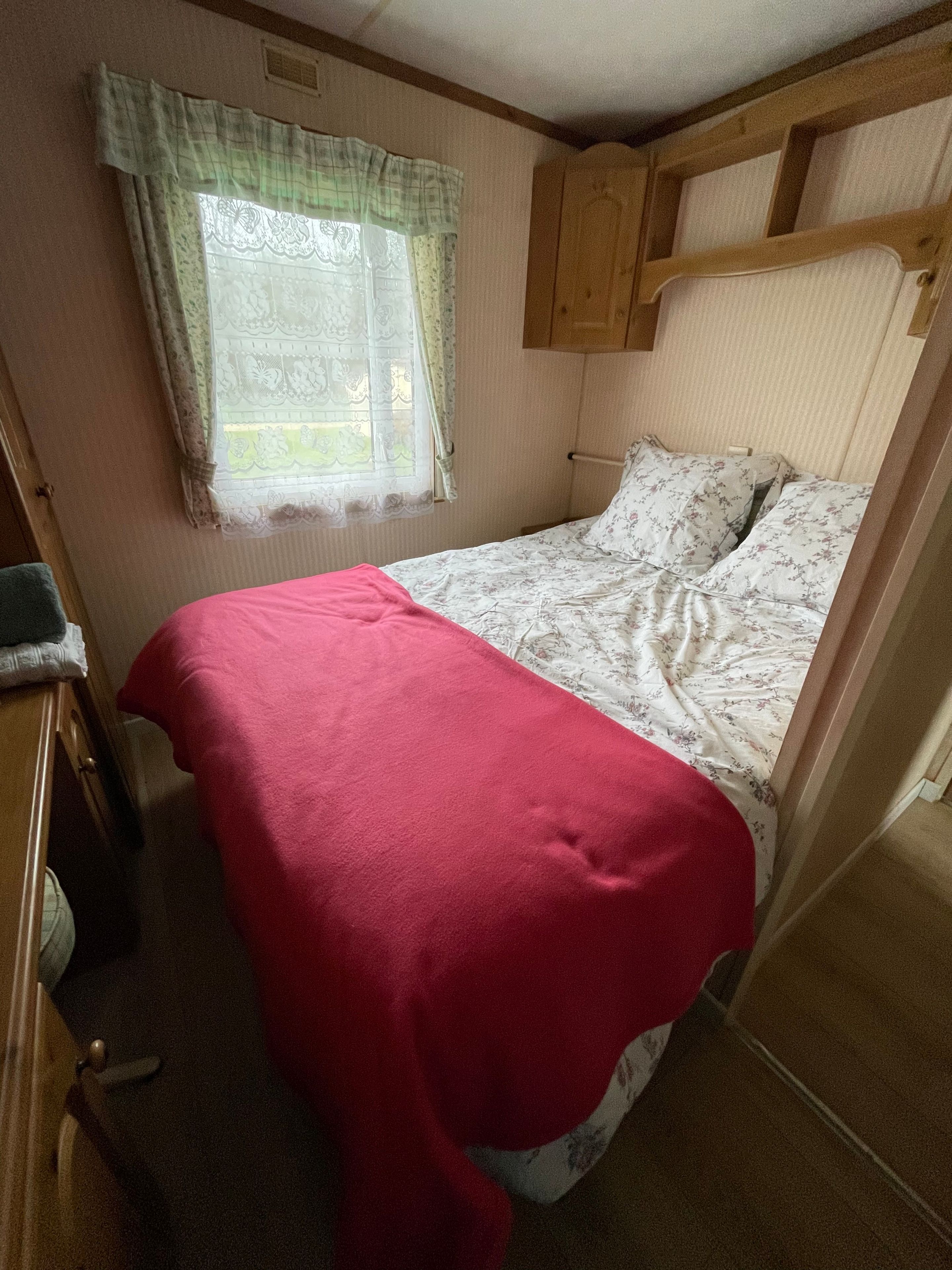 Un lit simple avec une couverture rouge, une fenêtre ornée de rideaux en dentelle et une tête de lit en bois.